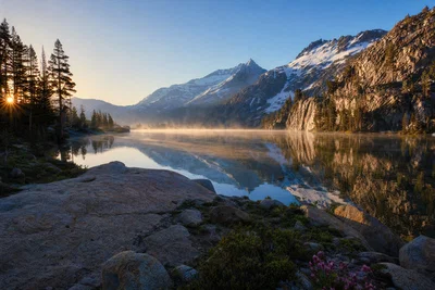 Alpine lake sunrise with mist, wide perspective, sharp foreground rocks, and snow peaks.