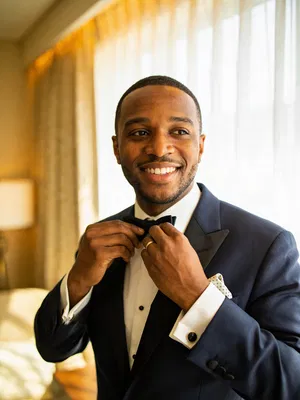 Black groom adjusting bow tie in hotel suite morning light