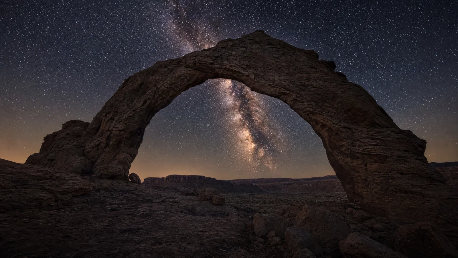 Desert rock arch framing the Milky Way with sharp stars, faint moonlight, and neutral night color.