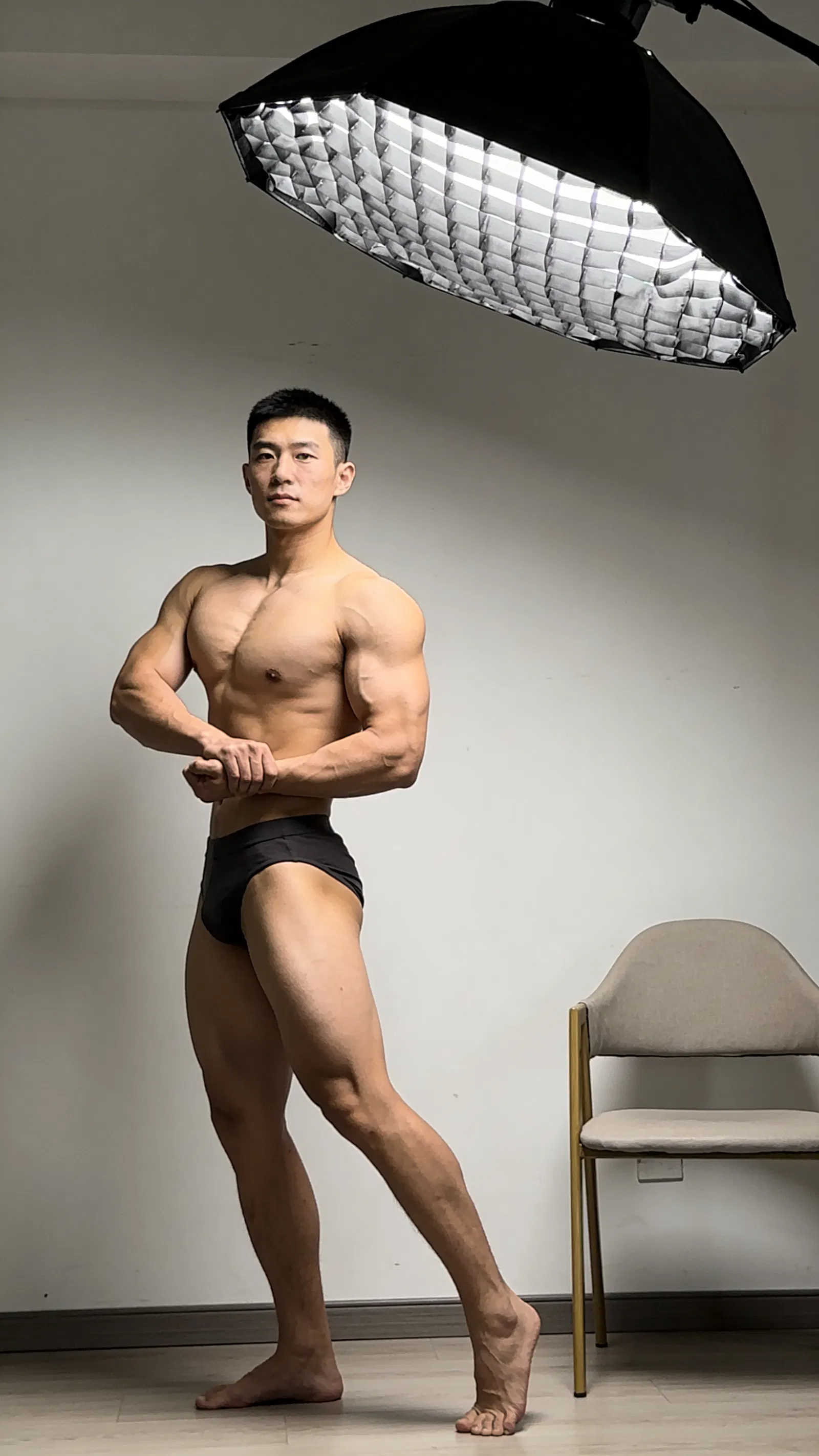 East Asian male bodybuilder side chest pose in simple home studio lighting