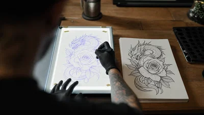 Hand tracing a tattoo stencil on a light table beside clean flash sketches.