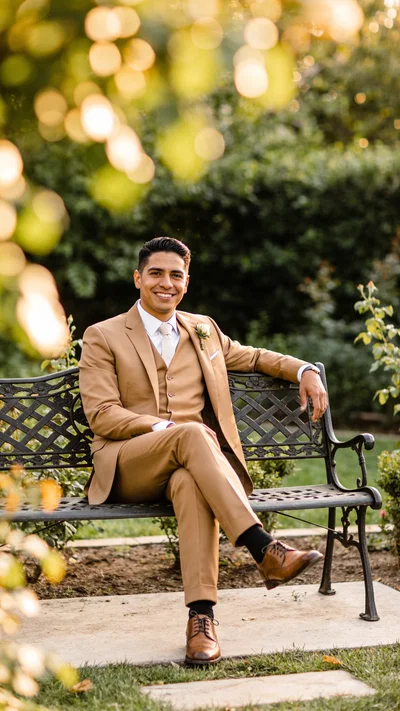 Hispanic groom in tan suit seated on garden bench with relaxed smile