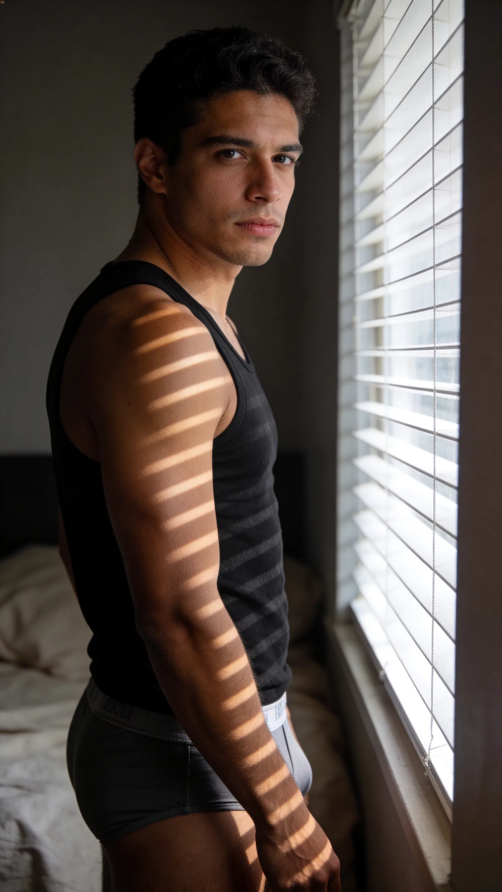 Hispanic man by window with blinds light pattern, men boudoir portrait mood