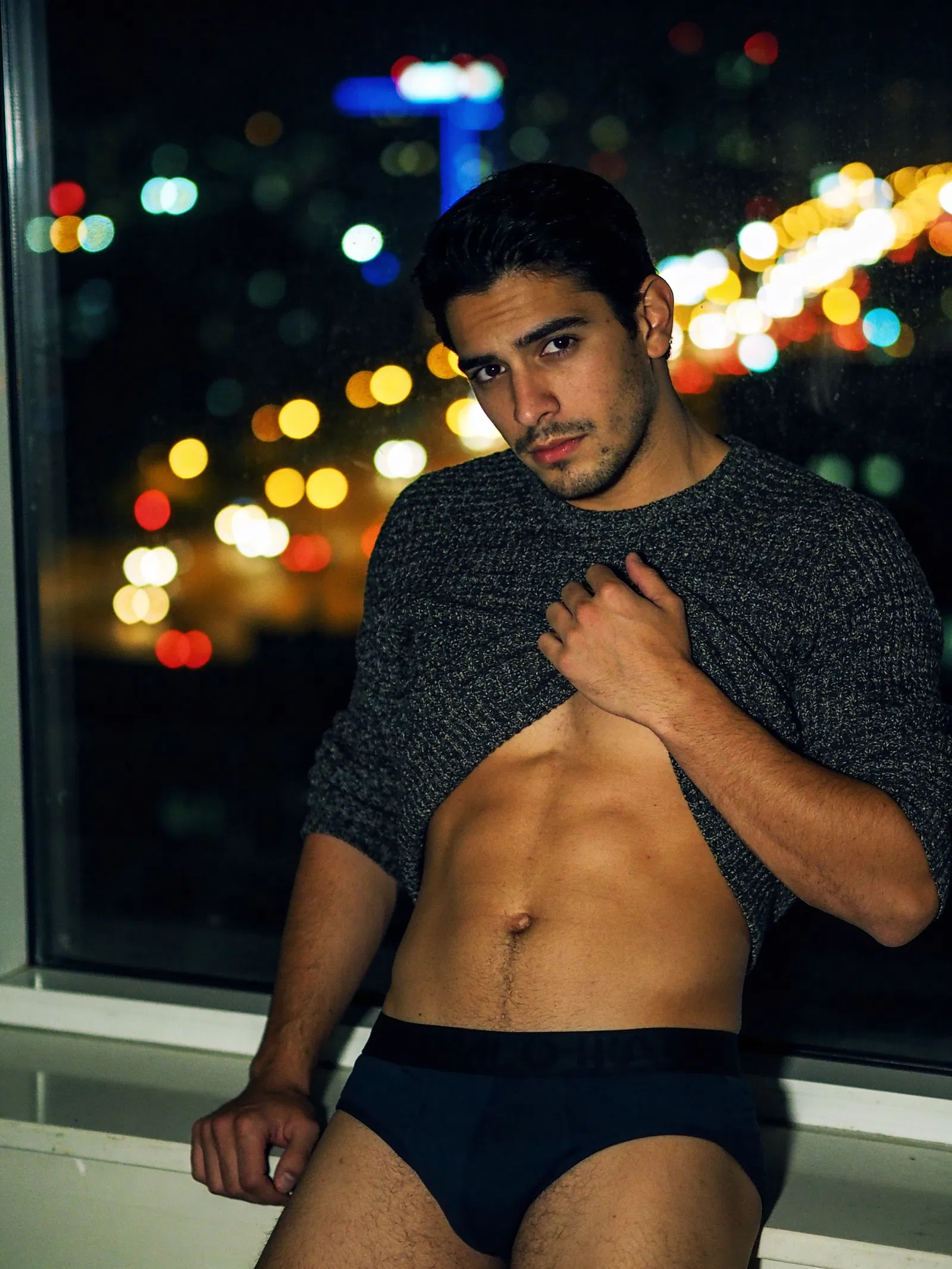 Hispanic man in moody window-lit male boudoir portrait with cinematic night bokeh