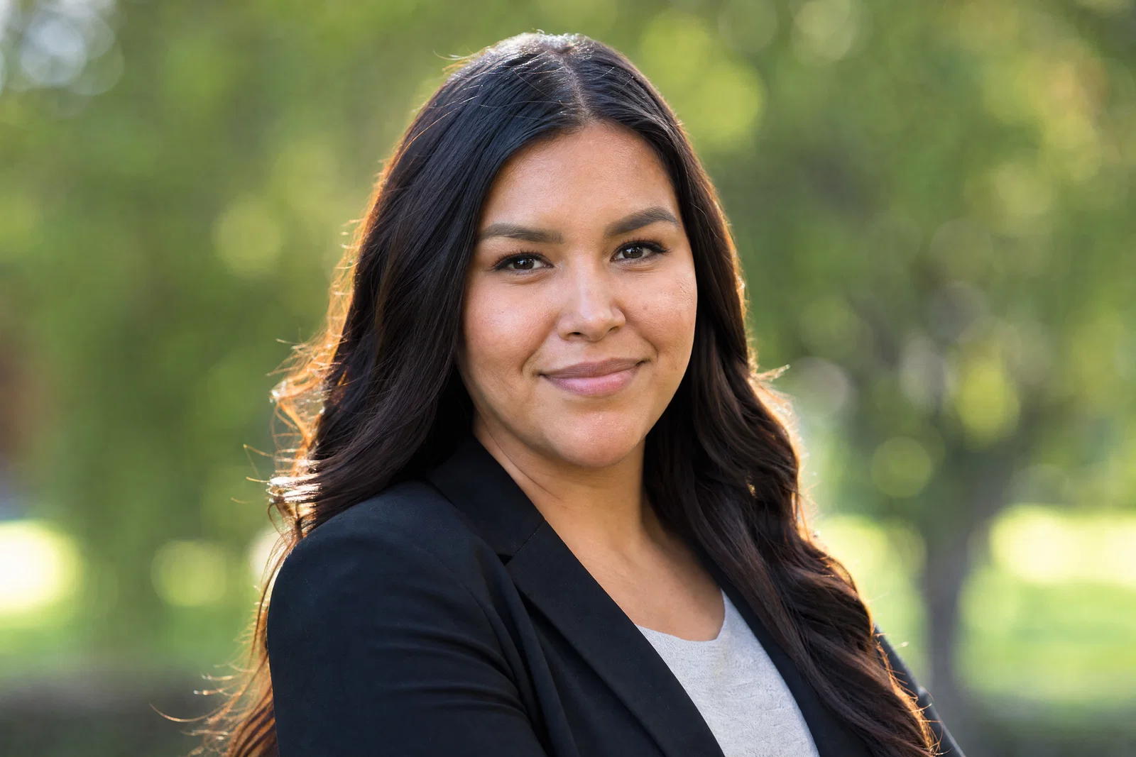 Indigenous woman in black blazer outdoors with softbox key, sun rim, and clean park bokeh.