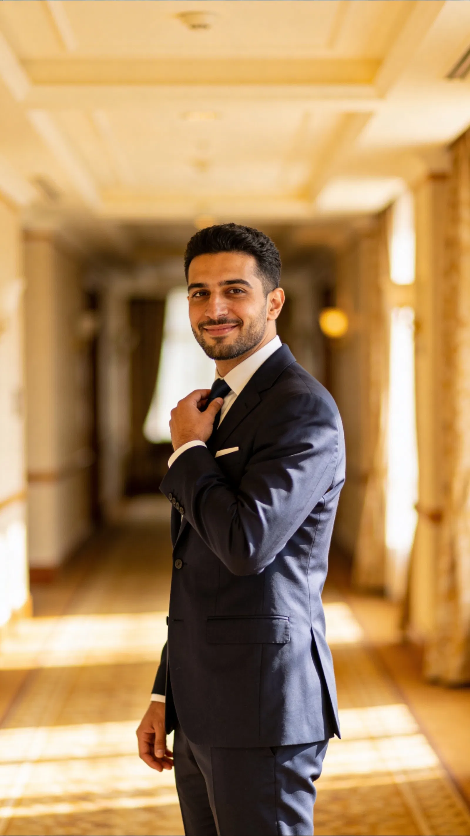 Middle Eastern groom adjusting tie in warm-lit hallway