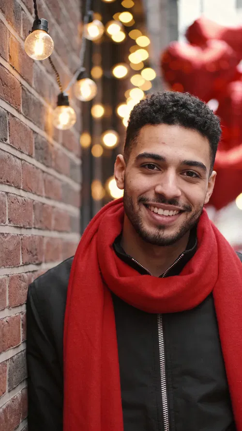 Middle Eastern man with red scarf leaning on brick wall, warm bokeh lights.