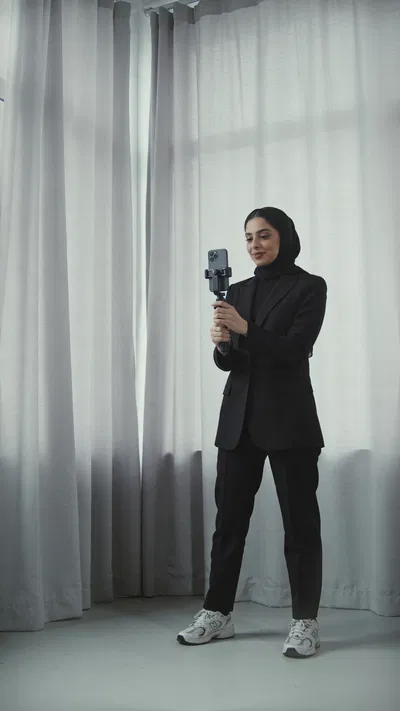 Middle Eastern woman in studio corner holding phone tripod, confident creator portrait