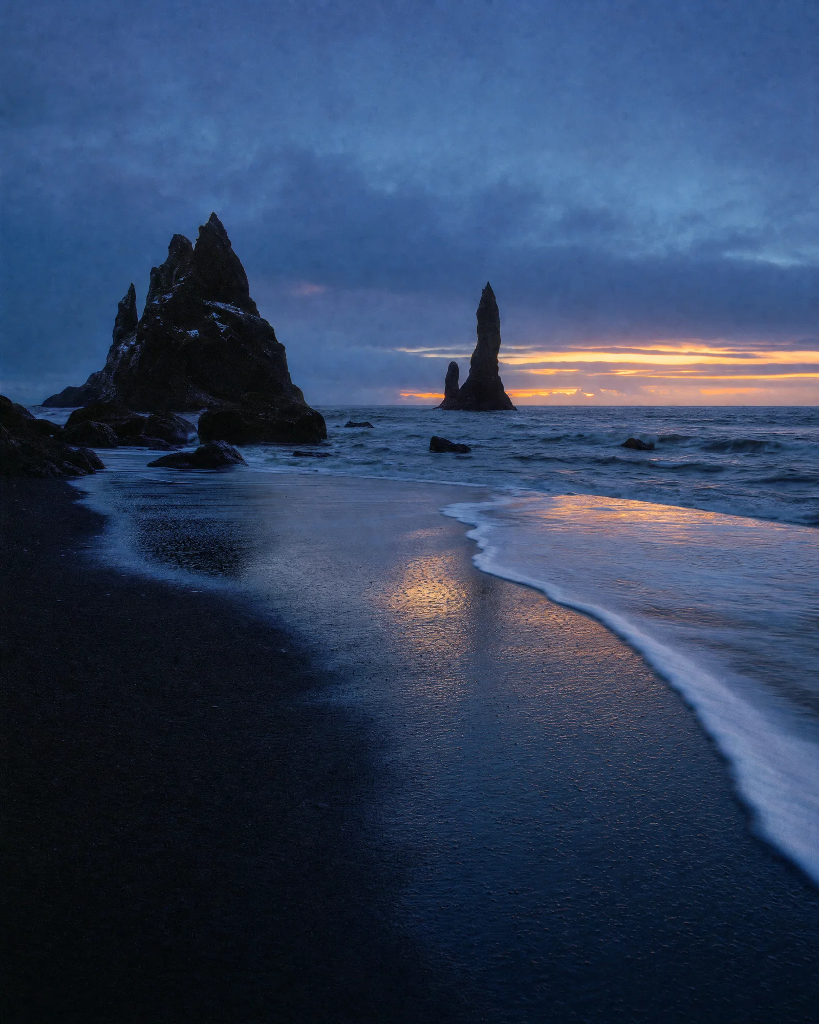 Vertical black sand beach at blue hour with basalt stacks, foam lines, and cool cyan shadows.