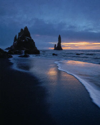 Vertical black sand beach at blue hour with basalt stacks, foam lines, and cool cyan shadows.