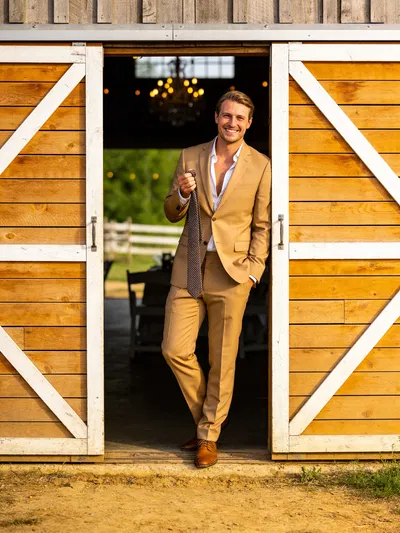White groom in tan suit at rustic venue doorway during prep
